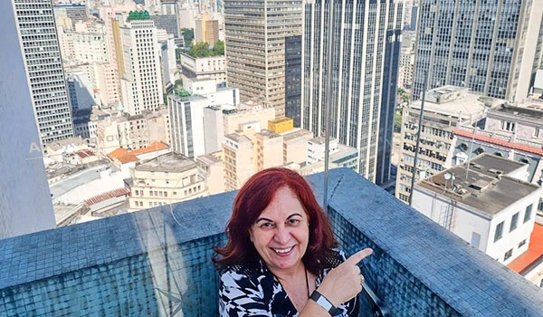 Deusa Rodrigues apontando para o lado em um terraço de prédio na sacada do Farol Santander na cidade de São Paulo, com vista de prédios altos ao fundo, destacando a palavra-chave sobre entretenimento.