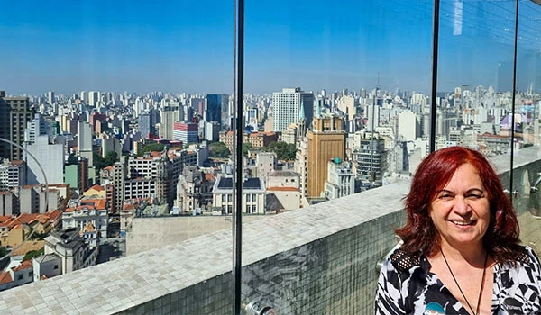 Deusa Rodrigues na sacada do Farol Santander com vista para a cidade de São Paulo ao fundo, refletida em uma grande janela de vidro.