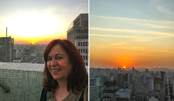 Deusa Rodrigues na sacada do Farol Santander com vista para o pôr do sol na cidade, destacando o cenário urbano ao entardecer.