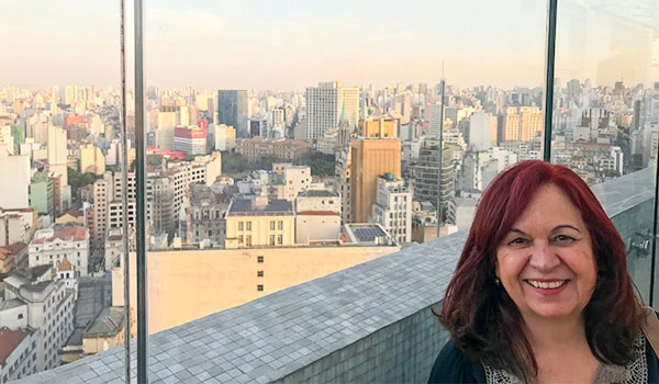 Deusa Rodrigues na sacada do Farol Santander com vista panorâmica de São Paulo ao fundo, dia ensolarado.