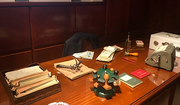 Imagem de uma mesa de escritório com livros, papelada, pasta, e objetos diversos, indicando um ambiente de trabalho ou escritório, com fundo de móveis de madeira escura.