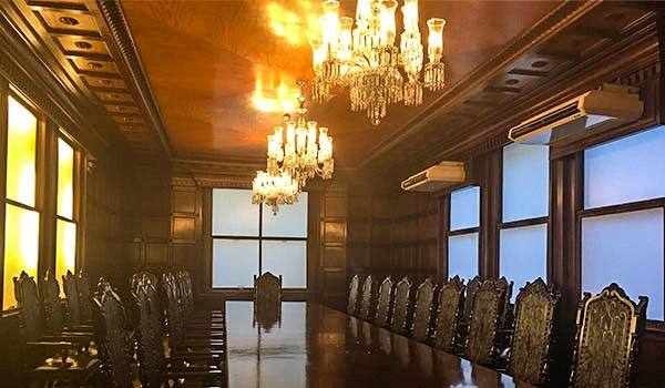 Imagem de uma sala de reunião ou conferência de luxo, com uma longa mesa de madeira, cadeiras elegantes e candelabros de cristal no teto, criando um ambiente sofisticado e elegante.