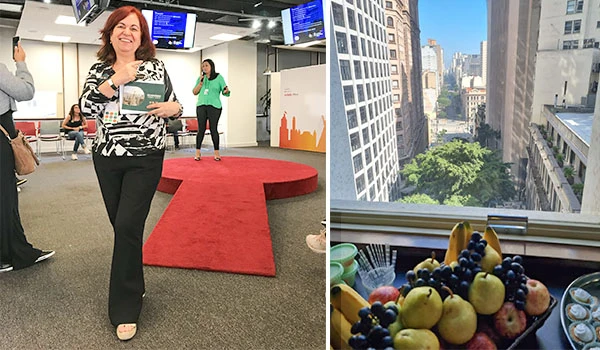 Deusa Rodrigues em evento corporativo com ambiente moderno, apresentando uma mesa com frutas variadas ao lado de uma vista de prédios altos na cidade
