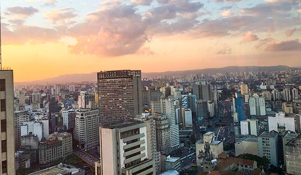 Vista aérea do centro de São Paulo ao entardecer, com o céu colorido de tons de rosa e laranja, apresentando diversos arranha-céus e prédios modernos