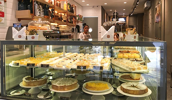 Imagem de uma cafeteria moderna com uma variedade de tortas e doces expostos em uma mesa de vidro, com uma atendente ao fundo e decoração elegante nas paredes.