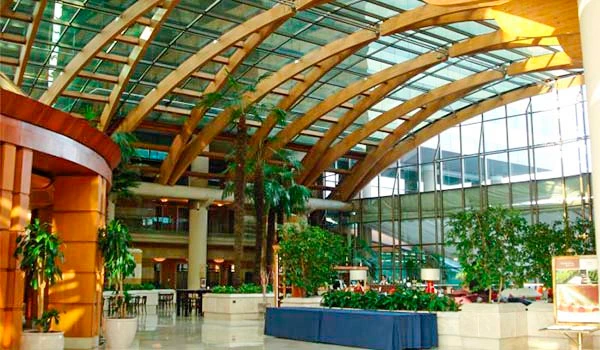 Interior de um shopping com teto de vidro arquitetônico, muitas plantas e espaço amplo com mesas de alimentação, ambiente natural e moderno.