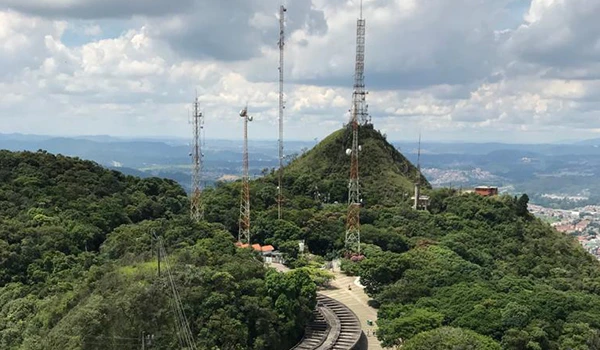 Paisagem de uma montanha com várias torres de transmissão de sinal em um cenário de natureza exuberante, céu nublado e áreas urbanas ao fundo, destacado por sua importância para a comunicação.