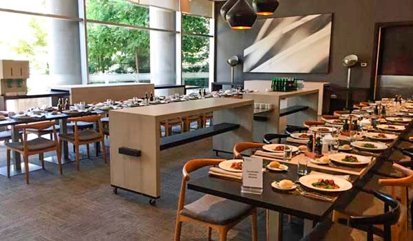 Ambiente de restaurante elegante com mesas bem arrumadas, pratos de comida e uma decoração moderna com janelas grandes que deixam entrar luz natural.