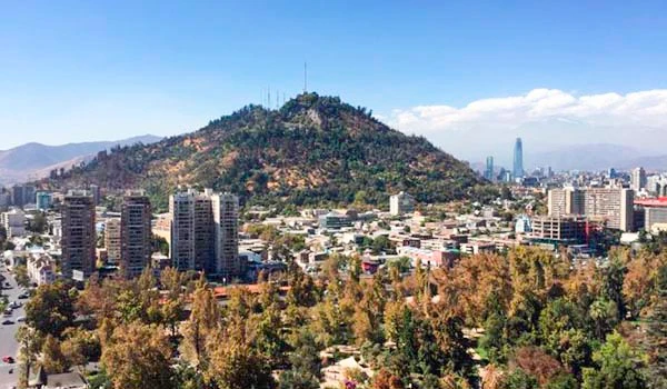 Vista panorâmica de uma cidade com um morro ao fundo, árvores coloridas na época do outono e edifícios altos, sob céu azul claro