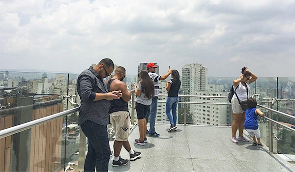 Grupo de pessoas aproveitando a vista em um terraço de prédio alto na cidade, com várias pessoas focadas em seus celulares e outras apreciando o panorama.