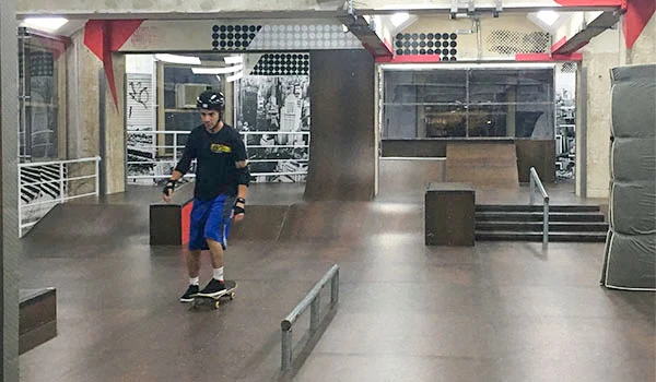 Imagem de um jovem praticando skate em um parque de skate coberto, com obstáculos como rampas e barras de tricks, em um ambiente urbano e moderno.