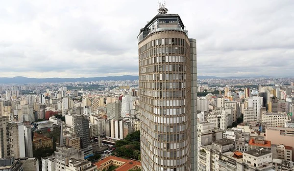 Vista panorâmica de um arranha-céu em São Paulo ao lado de uma vasta área urbana com muitos prédios e o céu nublado ao fundo.