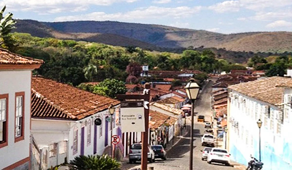 Vista de uma rua histórica em cidade de interior, com casas e fachadas antigas de telhado de cerâmica, vegetação ao redor e colinas ao fundo sob céu parcialmente nublado. Atrações do Planalto Central.