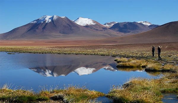 Paisagem de montanhas com neve ao fundo, refletidas em um lago tranquilo na planície. Dois visitantes observam a natureza na área.
