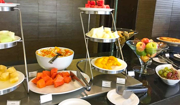 Mesa de buffet com saladas de frutas servidas em recipientes e tigelas, com melão, melancia e outros pedaços de frutas, além de copos com frutas ao fundo.