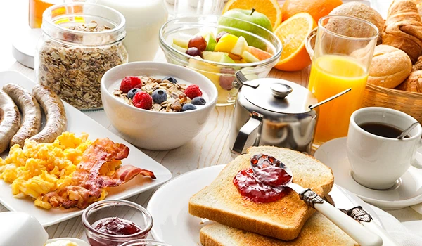 Café da manhã com frutas, iogurte, pão tostado, ovos e sucos em mesa posta, incluindo pães, bebidas quentes e frutas variadas em tigelas e copos.