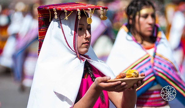 Mulher indígena em vestimenta tradicional participando de celebração cultural, usando um sombrero decorado e carregando um prato com alimentos tradicionais.
