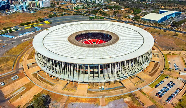 Vista aérea de um grande estádio de futebol moderno com arquibancadas circulares e telhado branco, campo central e entorno urbano ao redor.