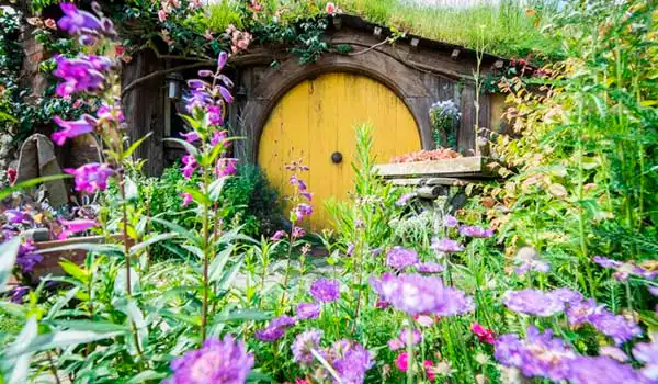 Jardim encantador com uma pequena casa com porta amarela, rodeada por flores coloridas, criando uma atmosfera mágica e acolhedora.