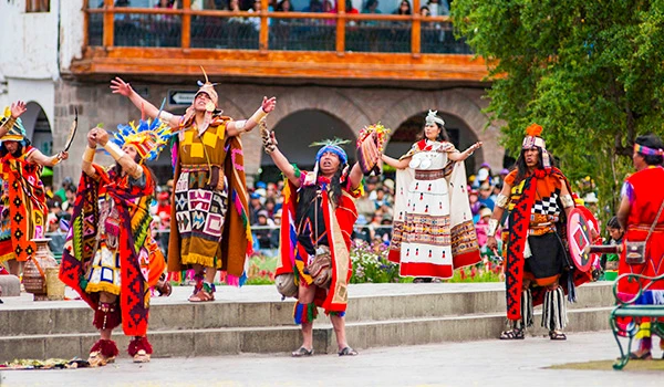 Dança tradicional indígena durante festival cultural ao ar livre, com participantes vestindo roupas coloridas e acessórios típicos, em cidade com árvores e arquitetura colonial ao fundo. Cultura e as danças da América do Sul.