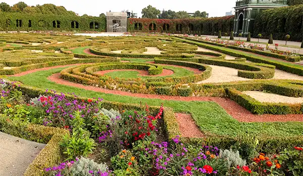 Jardim bem cuidado e decorativo com caminhos de terra, áreas de grama e flores coloridas, em um espaço ao ar livre com árvores e arquitetura histórica ao fundo.