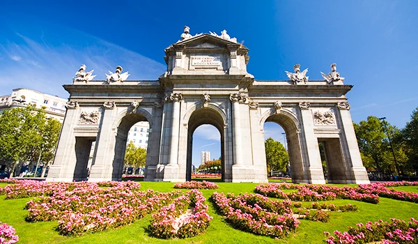 Imagem do Puerta de Alcala em Madri, Espanha, com detalhes arquitetônicos históricos e jardim de flores coloridas na frente, sob céu azul claro. Primavera na Europa.