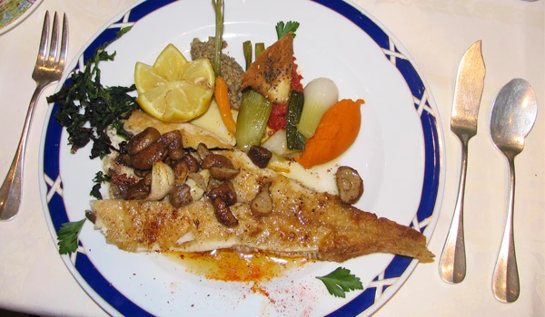 Prato de peixe grelhado com legumes variados como cenoura, quiabo, vagem, batata, e broto de couve, servido em um prato branco com detalhe azul.