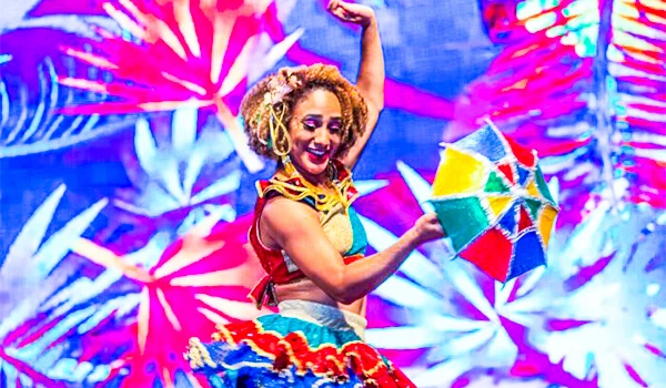 Mulher foliã dançando com sorriso no rosto ao som de samba, vestida com roupa típica colorida na festa folclórica brasileira, com fundo de cores vibrantes.