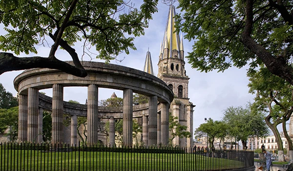 Foto da de uma praça com pliastras circulares e atrás uma igreja histórica com arquitetura imponente, torres altas e jardins bem cuidados ao redor, ideal para visitar na cidade. O que fazer em Guadalajara.