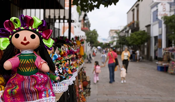 Feira de artesanato no México, com destaque para bonecas de tecido, roupas coloridas e pessoas caminhando na rua em dia ensolarado.