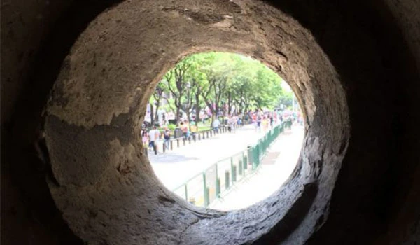 Vista de uma rua movimentada com árvores e pessoas, vista através de uma abertura em uma parede de pedra, criando um efeito de enquadramento natural. O que fazer em Guadalajara.