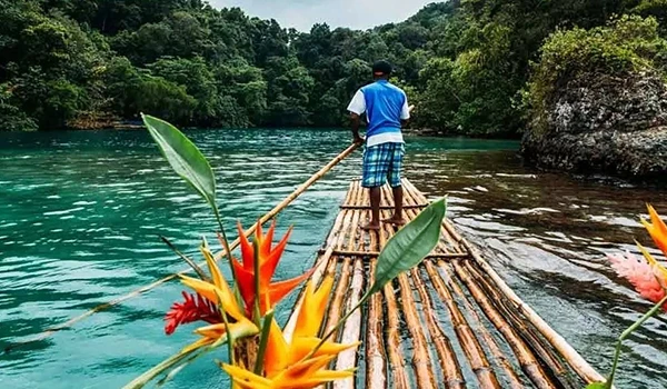 Homem navegando em uma balsa de madeira em um rio cercado por árvores verdes e natureza exuberante, cenário de turismo ecológico e aventura ao ar livre.