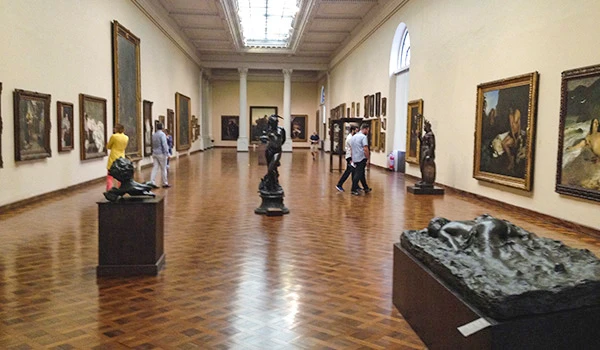 Interior de museu de arte com diversas obras e esculturas em exposição, visitantes apreciando as peças artísticas em uma galeria bem iluminada e espaçosa.