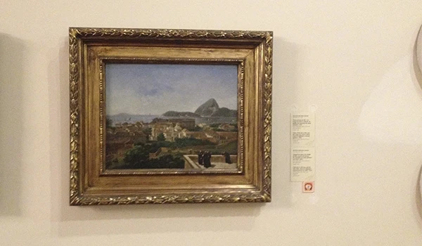 Quadro de paisagem com vista de montanhas, céu azul e elementos de cenário ao redor, exibido em uma galeria de arte com moldura dourada elaborada.