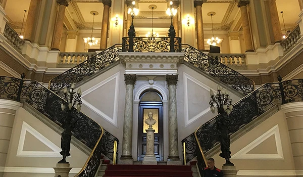 Imagem de uma grandiosa escadaria de um prédio histórico ou museu, com detalhes ornamentais, corrimãos de ferro trabalhado e iluminação elegante. Cultura no Rio de Janeiro.