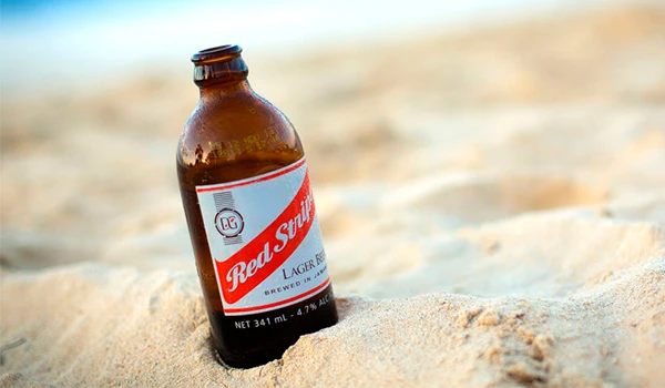 Imagem de uma garrafa de cerveja Red Stripe sobre a areia na praia, convidando para momentos de lazer e descanso à beira-mar.