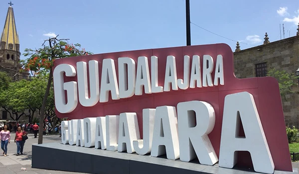 Letreiro comemorativo de Guadalajara, com destaque para o nome da cidade em letras grandes e brancas, localizado em um espaço ao ar livre com árvores e pessoas ao fundo.
