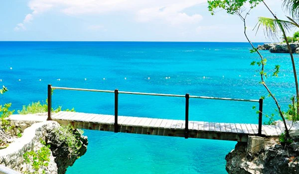 Imagem de uma ponte de madeira sobre águas cristalinas com litoral e céu azul ao fundo, cenário paradisíaco e tranquilo nas Jamaica praias.