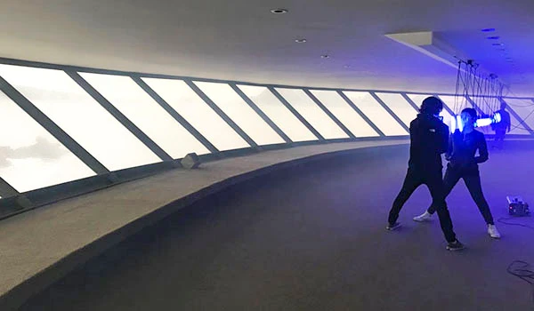 Imagem de um ambiente de estúdio com dois profissionais trabalhando com iluminação e câmeras, cenário futurista com janelas de vidro e iluminação azul.