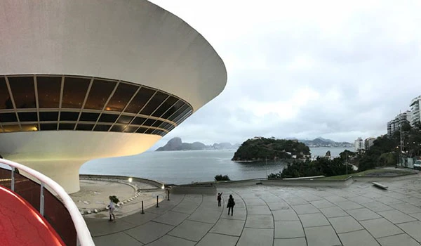 Visão panorâmica do Museu de Arte de Niterói, no Rio de Janeiro, com vista para a Baía de Guanabara ao fundo e céu nublado, destaque para a arquitetura moderna do museu.