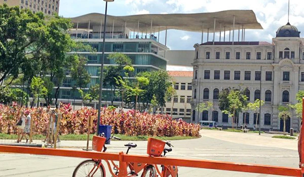 Imagem de praça movimentada com árvores, edifícios históricos e bikes estacionadas em um dia ensolarado, ideal para atividades ao ar livre. Cultura no Rio de Janeiro.