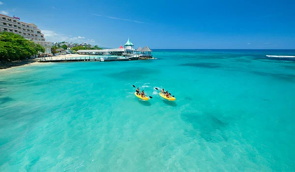 Praia da Jamaica paradisíaca com água cristalina, duas pessoas em caiaques e uma estrutura moderna ao fundo, ideal para relaxar e aproveitar o verão.