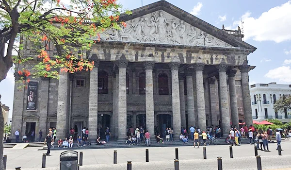 Fachada do Theatro Municipal de Guadalajara, um prédio clássico com colunas grandes e uma escultura no topo, cercado por visitantes e turistas em um dia ensolarado.