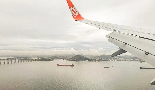 Imagem de uma vista aérea de um avião em voo sobre uma baía com navios e uma ponte, durante o dia nublado no Brasil.
