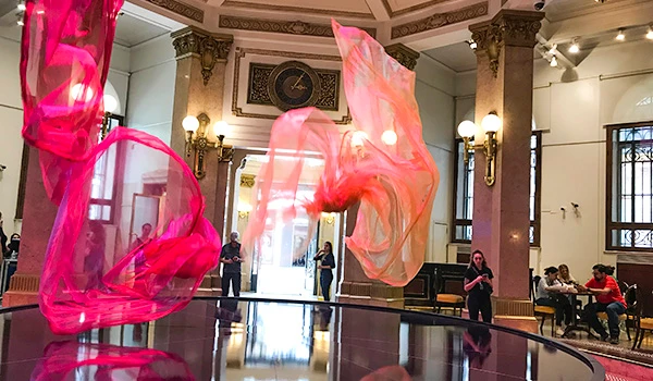 Instalação artística com tecidos de seda rosa fluindo sobre uma superfície refletora em um espaço amplo com detalhes clássicos na decoração, iluminação elegante e visitantes apreciando a arte.  Centro Cultural BB.