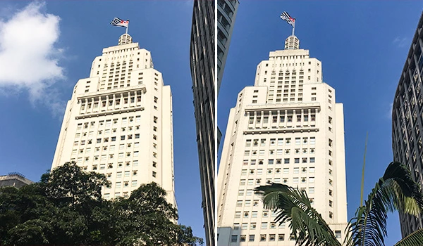 Imagem do edifício do Farol Santander no centro de uma cidade sob céu azul claro, com árvores na frente e uma bandeira de São Paulo no topo.