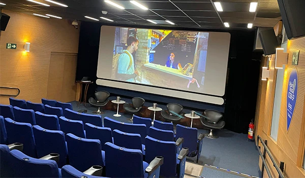 Sala de cinema com poltronas azuis, tela grande de projeção e duas pessoas assistindo a filme ou apresentação audiovisual.  Centro Cultural BB em São Paulo.