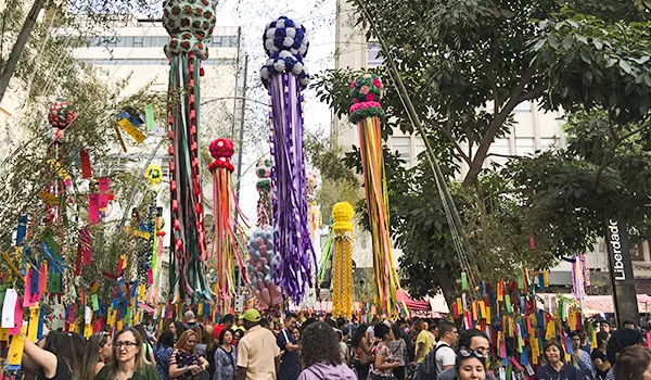 Multidão de pessoas participa de um festival ao ar livre em uma rua arborizada, decorada com grandes esculturas coloridas de flores e longas fitas pendentes, além de bandeirinhas e enfeites suspensos entre as árvores e os edifícios.