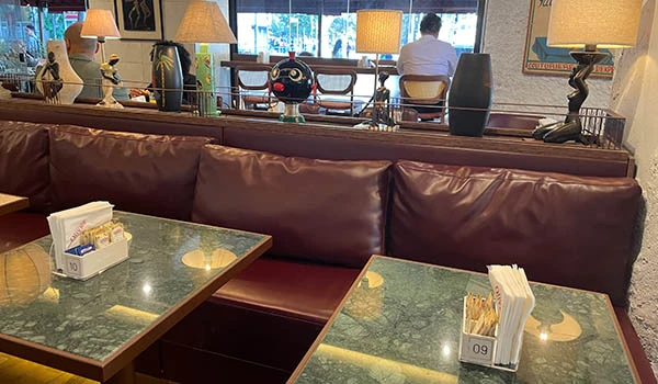 Imagem de um restaurante sofisticado com poltrona de couro vinho, mesas de vidro com guardanapos e utensílios, e decoração elegante com portas de vidro ao fundo.