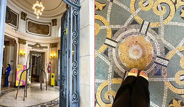 Imagem do interior de uma fachada de edifício clássico, com detalhes decorativos dourados e entrada com portas de ferro ornamentadas, ao lado de um piso com mosaico em forma de mandala, onde uma pessoa está de pé.  Centro Cultural BB.